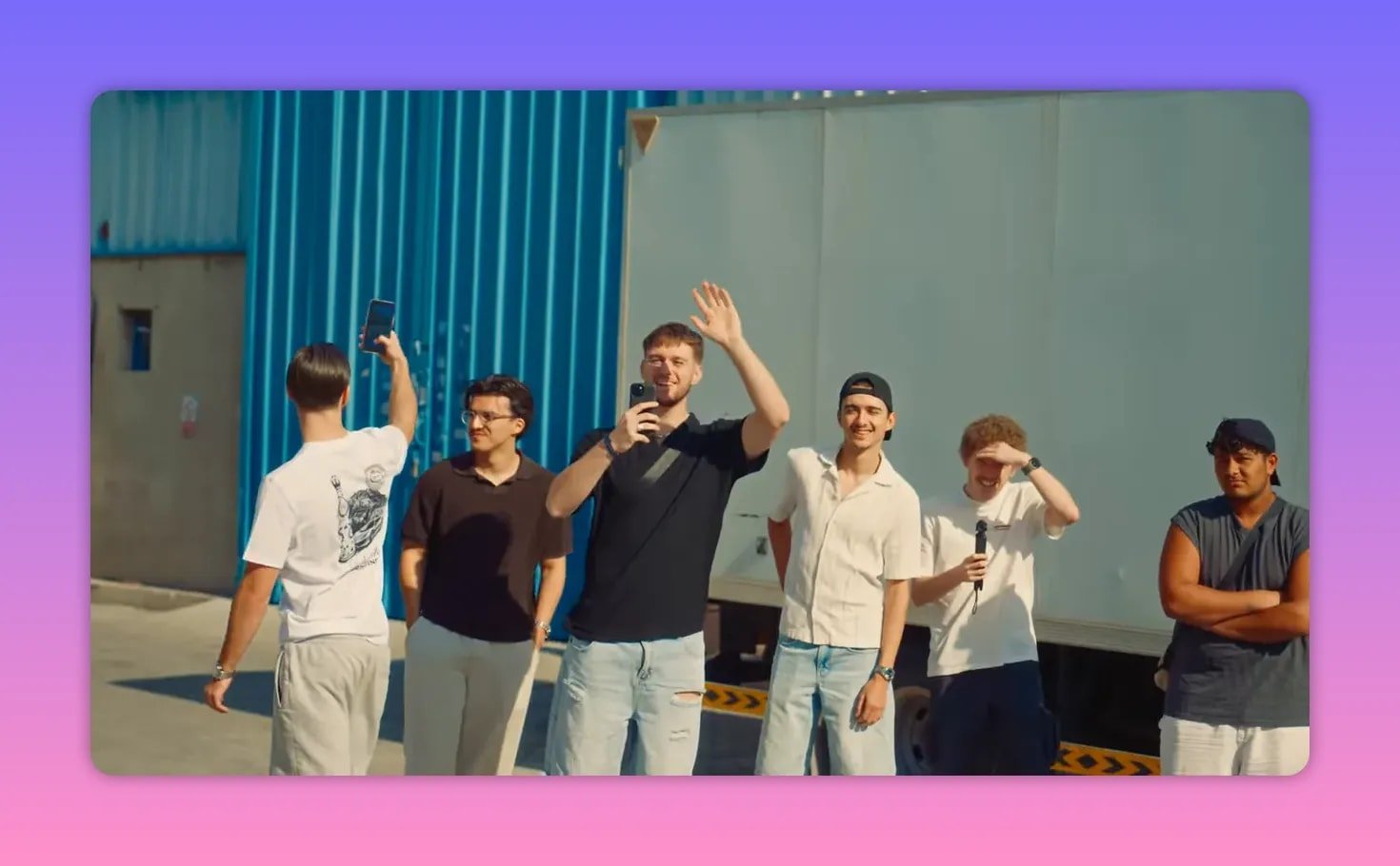 Groupe de jeunes hommes souriants, certains filmant et saluant, devant un entrepôt